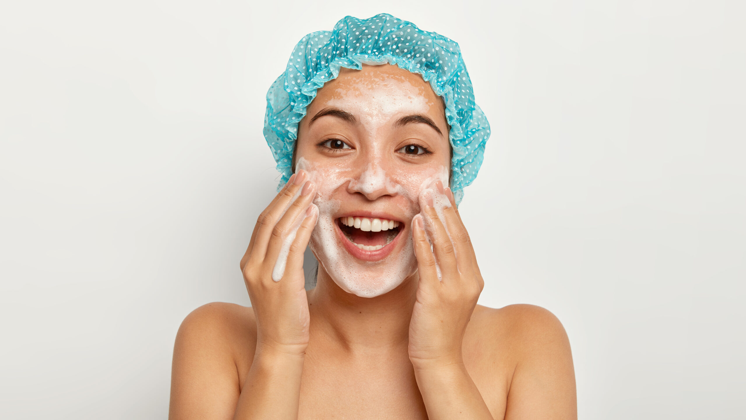 Photo of lovely female model with happy expression, washes face with foaming cleanser, wears wateproof showercap, pampers skin, stands shirtless, looks straightly at camera. Facial treatment